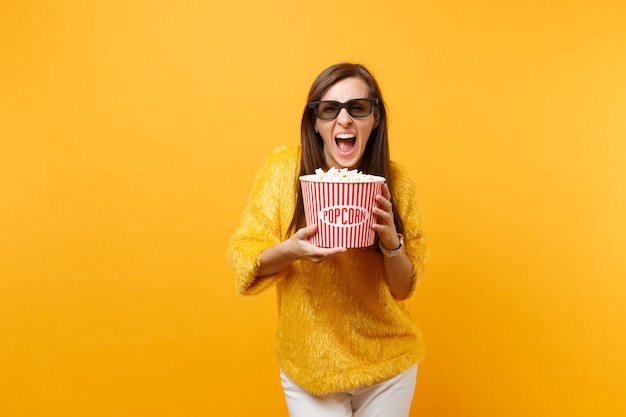 Chica joven loca en gafas imax 3d viendo la película gritando sosteniendo un cubo de palomitas de maíz aislado sobre fondo amarillo brillante. Personas sinceras emociones en el concepto de estilo de vida de cine. Área de publicidad.