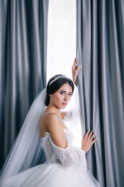 Chica joven en un lujoso vestido de novia con un velo largo cerca de la ventana en el apartamento Mujer con cabello negro con un peinado de novia mañana novia en una habitación moderna