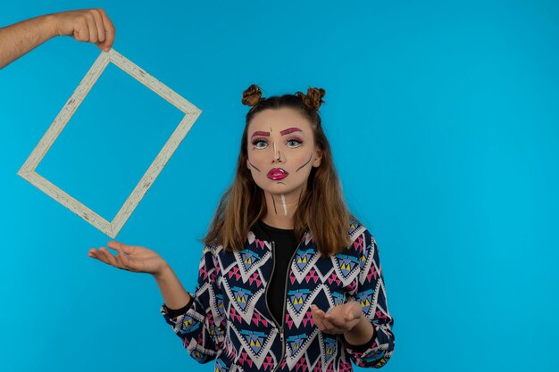 Chica joven con maquillaje de fantasía con marco vacío sobre fondo azul. foto de alta calidad