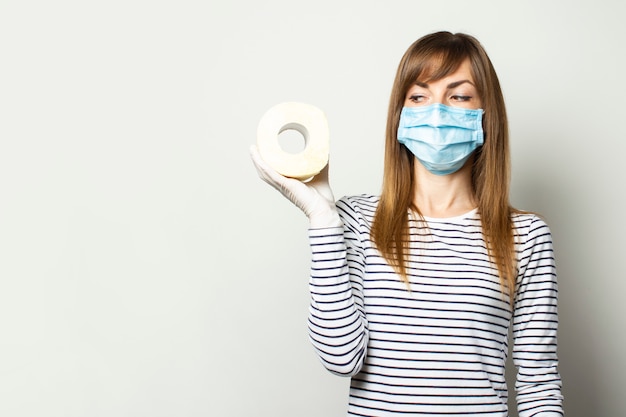 Chica joven en una máscara médica y guantes médicos sosteniendo un rollo de papel higiénico y mirarlo en una pared de luz. Concepto de cuarentena, escasez, pánico