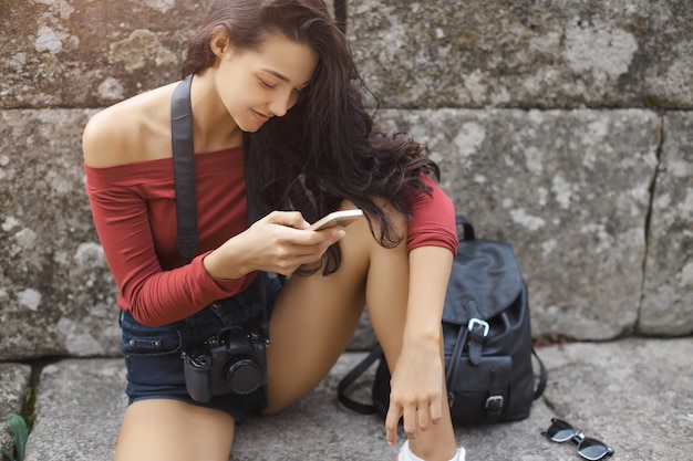 Chica joven con mochila para smartphone y cámara al aire libre. Feliz mujer vestida con ropa casual