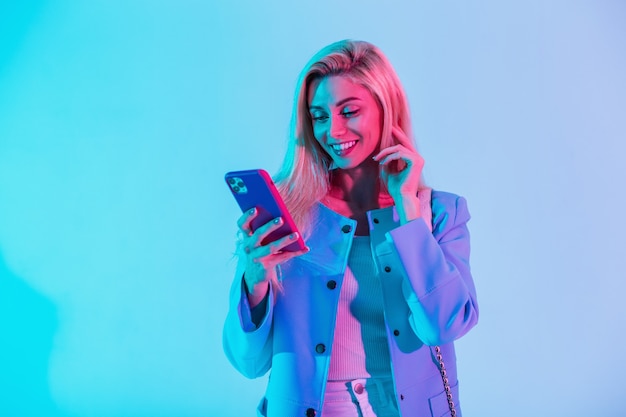 Chica joven de moda feliz con una sonrisa de belleza en ropa de negocios elegante de moda con una chaqueta sosteniendo un teléfono inteligente y escribiendo un mensaje en una luz de neón rosa multicolor en el estudio
