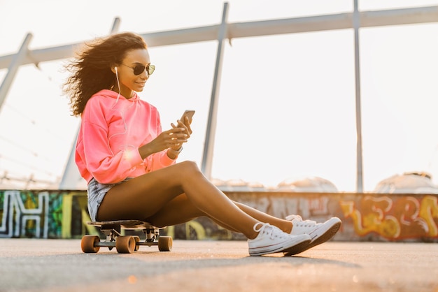 Chica joven monopatín usando teléfono y auriculares al aire libre