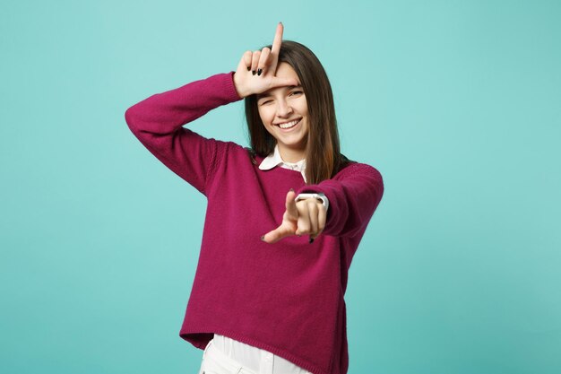 Chica joven morena con ropa informal posando aislada en un retrato de estudio de fondo de pared azul. Concepto de estilo de vida de las personas. Simulacros de espacio de copia. mostrando el gesto de perdedor apuntando con el dedo índice a la cámara