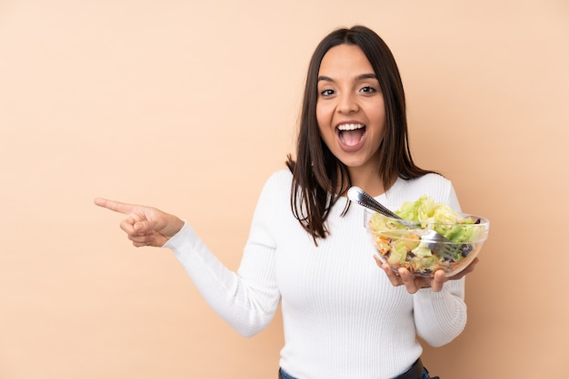 Chica joven morena sosteniendo una ensalada aislado sorprendido y apuntando con el dedo hacia un lado