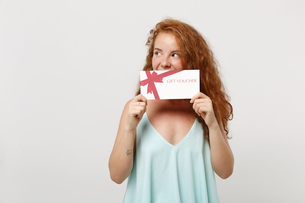 Chica joven mujer bonita pelirroja en ropa ligera casual posando aislada sobre fondo de pared blanca. Concepto de estilo de vida de personas. Simulacros de espacio de copia. Cubriendo la boca con certificado de regalo, mirando a un lado.