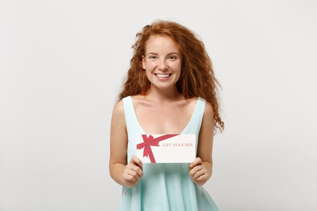 Chica joven pelirroja sonriente en ropa ligera casual posando aislada sobre fondo de pared blanca, retrato de estudio. Concepto de estilo de vida de personas. Simulacros de espacio de copia. Sosteniendo en las manos certificado de regalo.