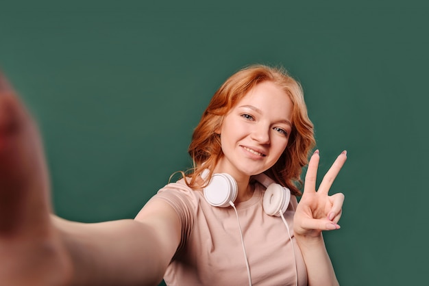 Chica joven pelirroja toma una selfie en primera persona y muestra un signo victoriano, de pie aislado en una pared verde con lugar para el texto.