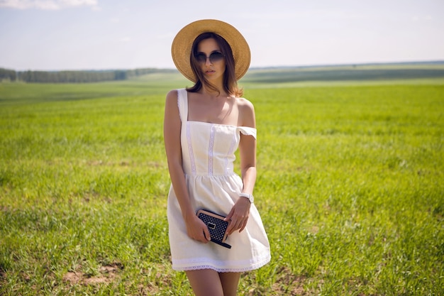 Chica joven con pelo largo y negro se encuentra en el sitio con vestido blanco y sombrero de paja de ropa está en el campo verde con hierba y sonrisas, verano.