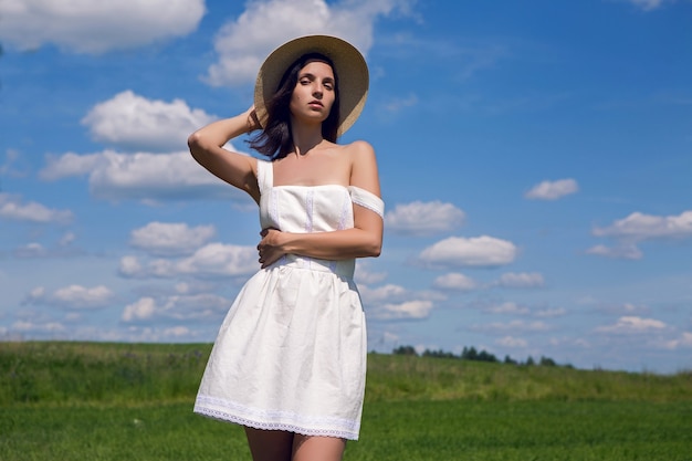 Chica joven con pelo largo y negro se encuentra en el sitio con vestido blanco y sombrero de paja de ropa está en el campo verde con hierba y sonrisas, verano.