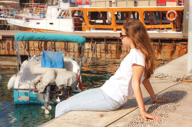 La chica joven con el pelo largo se sienta en el embarcadero en el puerto del yate y mira el mar azul