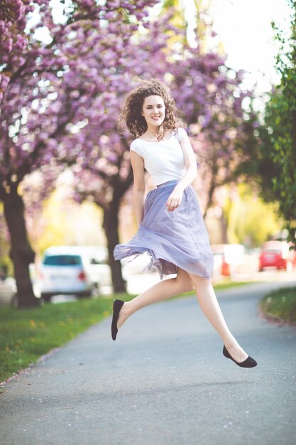 Chica joven con el pelo rizado en una camiseta blanca y falda violeta bailando y saltando bajo los árboles florecientes de sakura