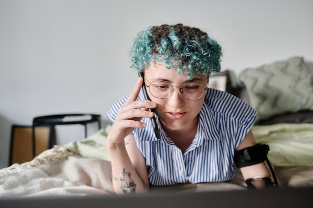 Chica joven con el pelo rizado teñido conversando por teléfono móvil mientras usa la computadora portátil