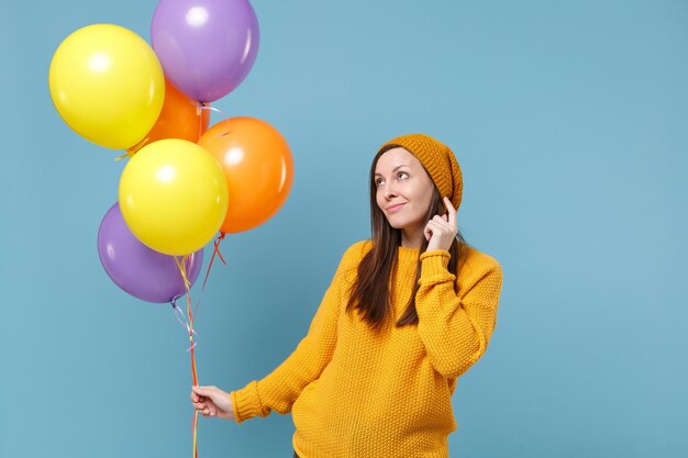 Chica joven pensativa en suéter y sombrero posando aislado sobre fondo azul. Concepto de emociones de la gente de la fiesta de cumpleaños. Simulacros de espacio de copia. Celebrando sostener globos de aire coloridos mirando hacia arriba.