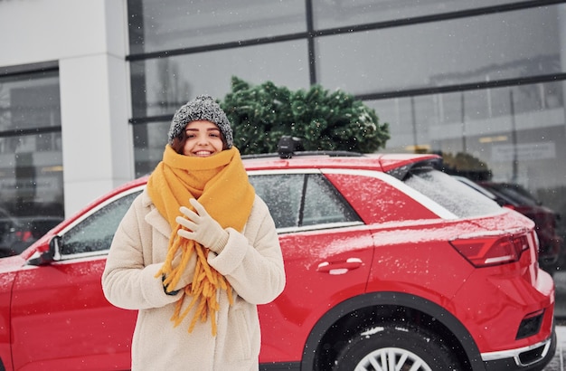 Chica joven positiva de pie cerca del coche con árbol de Navidad verde en la parte superior.