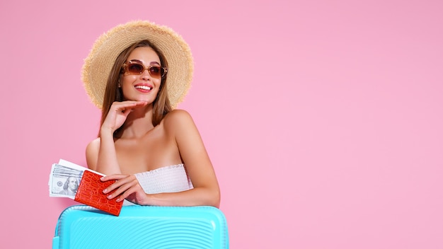 Chica joven positiva con sombrero de paja y gafas de sol con boletos y maleta fondo rosa estudio smi ...