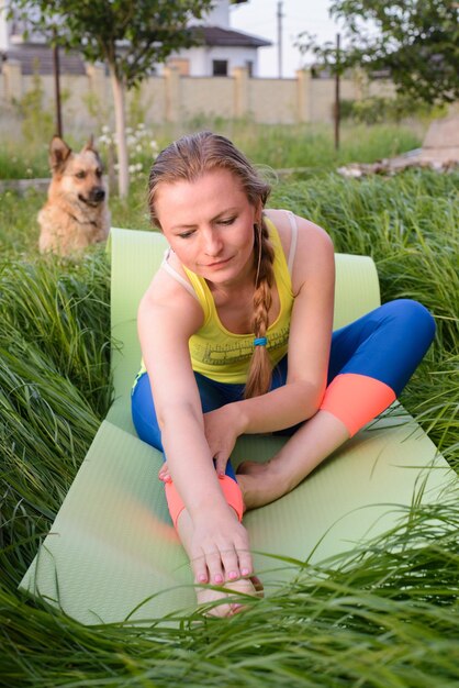 Chica joven que hace yoga en el patio trasero y un perro que se sienta en el fondo