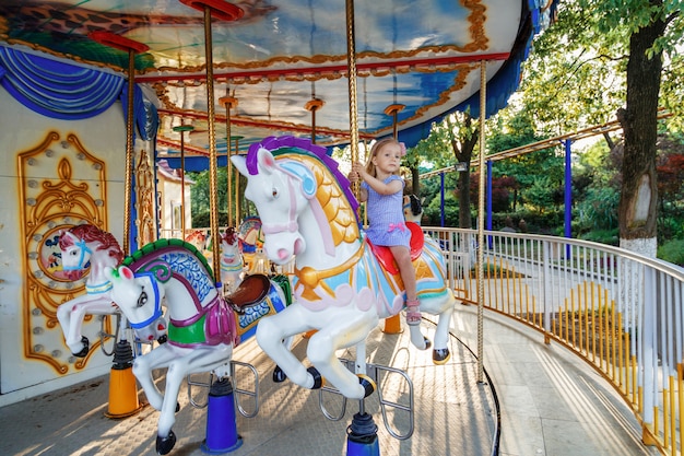 Chica joven que monta en caballo del parque de atracciones en el paseo de la diversión del carrusel en el parque de los parques de atracciones al aire libre