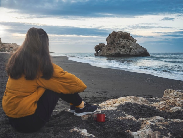 Chica joven que se relaja en una playa al amanecer con una taza de café En foco en el fondo