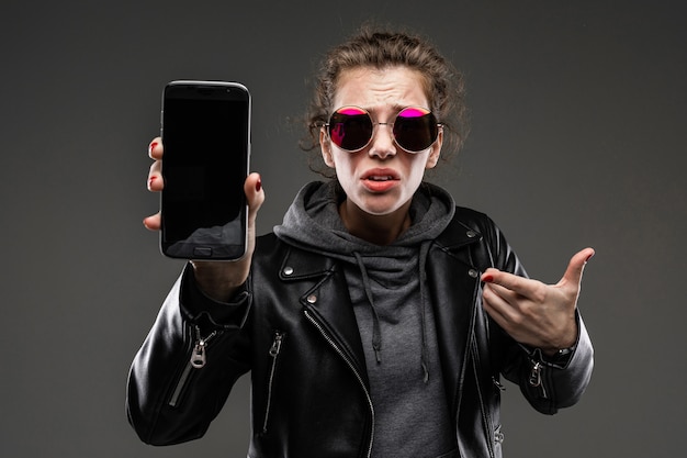 Chica joven con rasgos faciales ásperos, cabello castaño acariciado, manicura brillante, bicicleta gris, chaqueta negra, se muestra en el teléfono y no está contenta