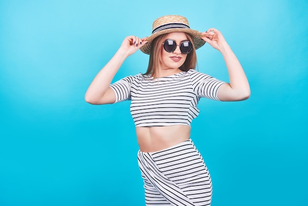 Chica joven con rayas blancas y negras, sombrero, gafas de sol, boca abierta emocionalmente sobre un fondo azul brillante con un cuerpo perfecto. Aislado. Tiro del estudio.