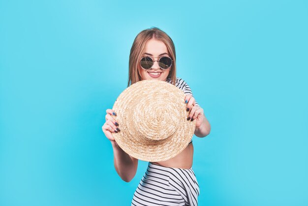 Chica joven con rayas blancas y negras, sombrero, gafas de sol, boca abierta emocionalmente sobre un fondo azul brillante con un cuerpo perfecto. Aislado. Tiro del estudio.