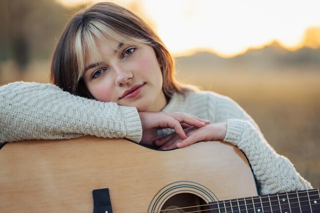 Foto chica joven relajada en suéter blanco sentada con la guitarra en la naturaleza puesta de sol en el fondo