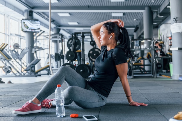 Chica joven en ropa deportiva tomando un descanso con comida y agua está en el gimnasio durante el día.