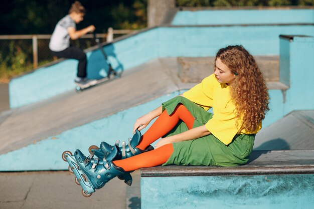 Chica joven en ropa verde y amarilla y medias naranjas con peinado rizado patinaje sobre ruedas sentado en skate park