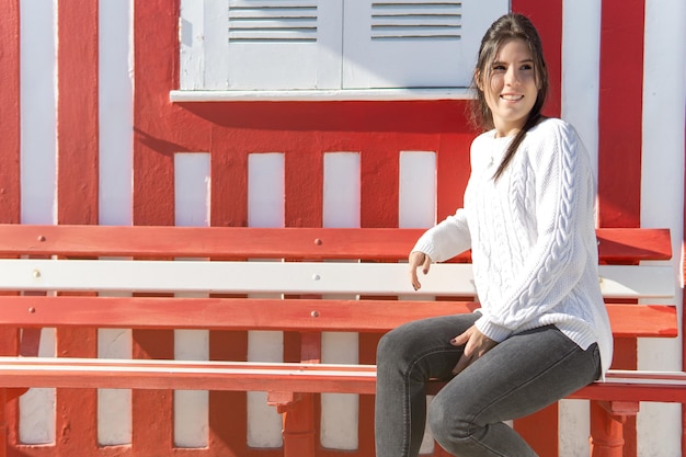 Chica joven sentada en un banco con una pared de líneas rojas y blancas en el fondo