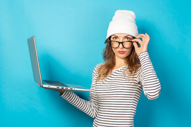 Chica joven seria con una computadora portátil y un sombrero blanco, se ve desde debajo de las gafas en azul