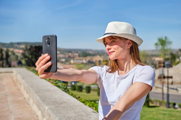 Chica joven con un sombrero blanco sonriendo sentada en un jardín tomando un selfie con su teléfono inteligente