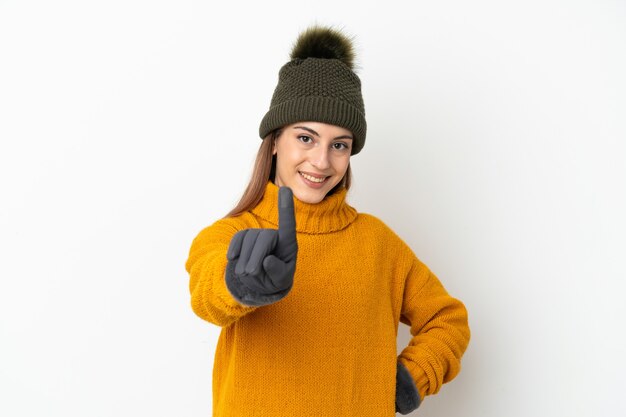 Chica joven con sombrero de invierno aislado en la pared blanca mostrando y levantando un dedo