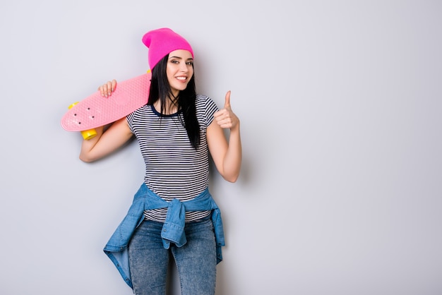 Chica joven con sombrero rosa y patineta sobre su hombro sonriendo y mostrando el pulgar hacia arriba