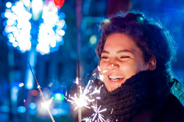 Chica joven sonriente celebración de sparkler en su mano. Sonriente adolescente en la calle en la noche con sparklers