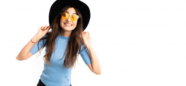 Chica joven con una sonrisa encantadora, cabello castaño largo y ondulado, maquillaje hermoso, jersey azul, jeans negros, boina gris, anteojos, sombreros negros y sonrisas