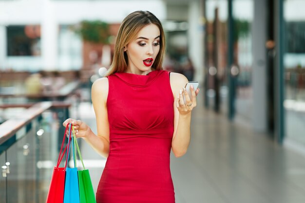 Chica joven sorprendida con cabello castaño claro y labios rojos de pie con bolsas de colores, sosteniendo el teléfono móvil, concepto de compras, retrato.