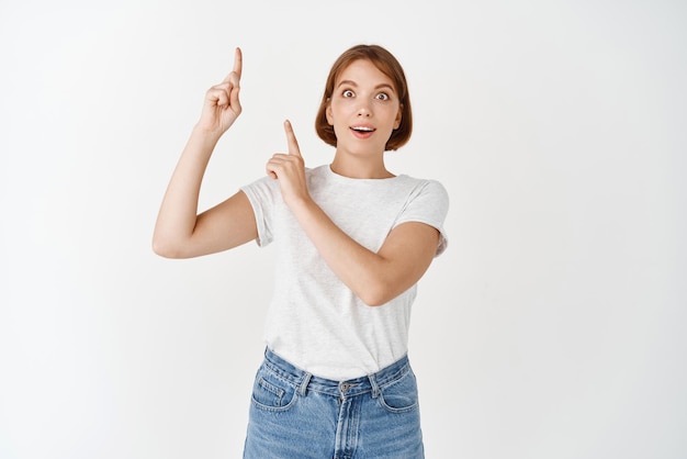 Chica joven sorprendida y emocionada en camiseta apuntando con los dedos hacia arriba jadeando asombrada y sonriendo mostrando una promoción genial de pie sobre fondo blanco