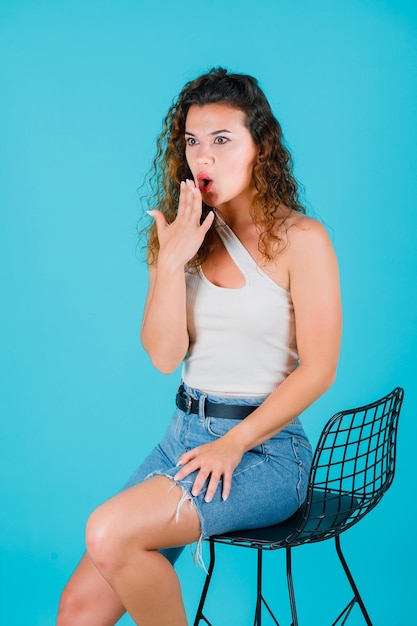 Chica joven sorprendida está sosteniendo la mano en la boca sobre fondo azul.