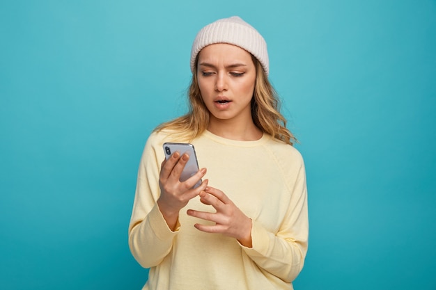 Chica joven sorprendida con sombrero de invierno sosteniendo y mirando el teléfono móvil