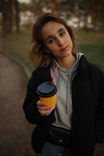 La chica joven sostiene un vaso de café en la mano. chica bebe un café de un vaso de papel en la calle