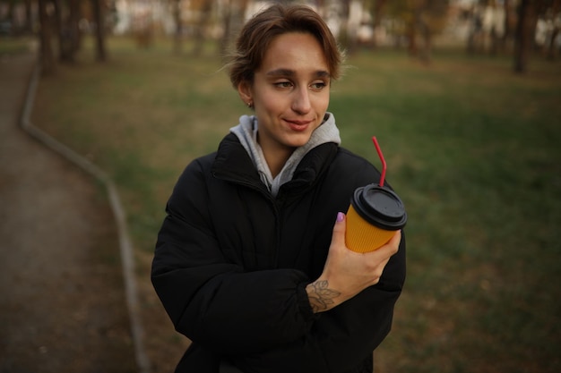 La chica joven sostiene un vaso de café en la mano. chica bebe un café de un vaso de papel en la calle