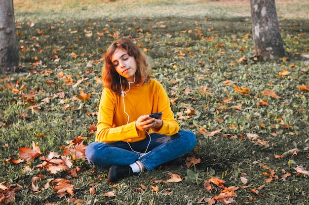 Chica joven en suéter amarillo escuchando música con auriculares sentados en la hierba de otoño