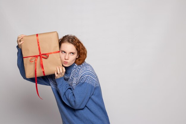 Chica joven en un suéter sostiene un regalo sobre un fondo blanco.