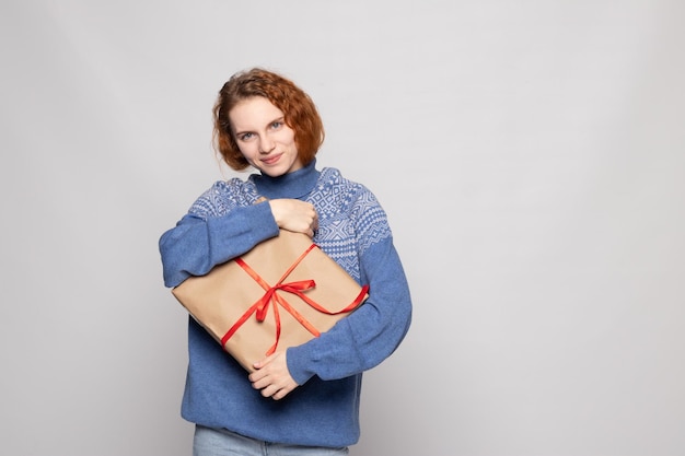 Chica joven en un suéter sostiene un regalo sobre un fondo blanco.