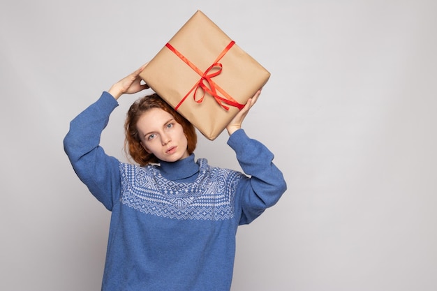 Chica joven en un suéter sostiene un regalo sobre un fondo blanco.