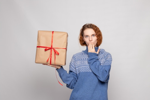 Chica joven en un suéter sostiene un regalo sobre un fondo blanco.