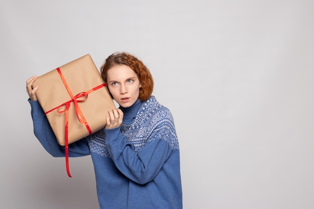 Chica joven en un suéter sostiene un regalo sobre un fondo blanco.