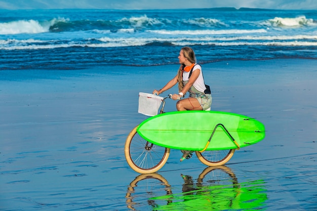 Chica joven con tabla de surf y bicicleta en la playa