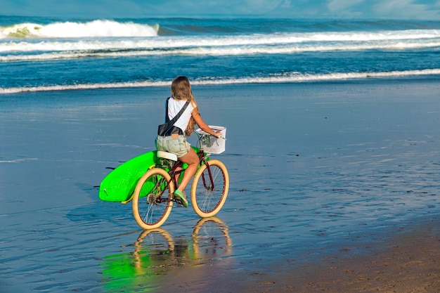 Chica joven con tabla de surf y bicicleta en la playa